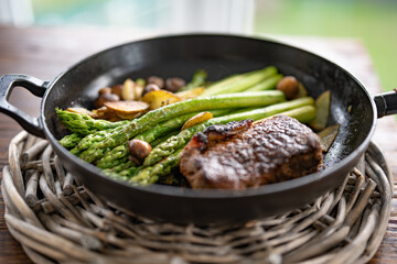 Crispy roasted beef steak with green asparagus, mushrooms, and potato slices in a cast iron pan. Close-up with shallow depth of field.