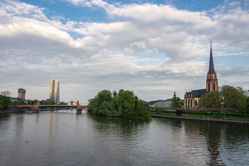 Frankfurt's Main River flows past the Dreikonigskirche spire, a historic stone bridge, and the modern ECB tower under a partly cloudy sky.