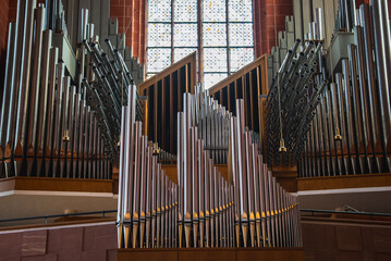 A grand pipe organ with intricate metal pipes stands before a large stained glass window. Red brick walls and wooden details highlight the historic interior.