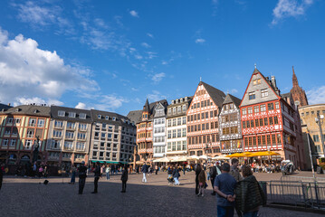 Obraz premium Romerberg square in Frankfurt features colorful half timbered buildings, the Frankfurt Cathedral spire, and people enjoying the lively open space.
