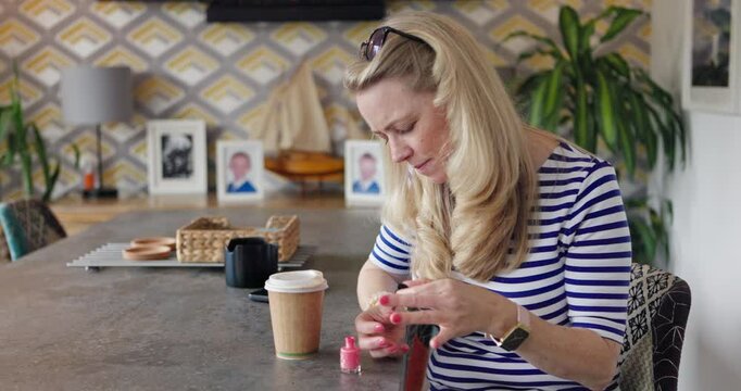 Woman, blow nail polish and home with manicure, makeup or cosmetics with coffee, self care and desk. Person, grooming and painting fingernails with style for hand, relax and color on table in house