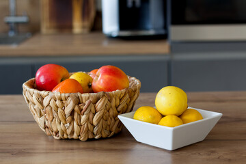 A woven basket holds apples, oranges, and lemons, while a white square bowl contains several lemons on a wooden countertop in a kitchen setting