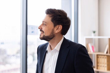 Young adult Caucasian male in professional suit smiling while standing indoors near window. Business, confidence, and focus depicted in modern workplace setting.