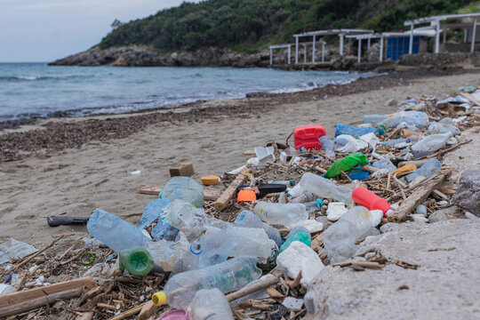 Large amount of plastic bottles, broken objects, and wooden debris covering sandy beach coastline. Ocean pollution and environmental degradation crisis.