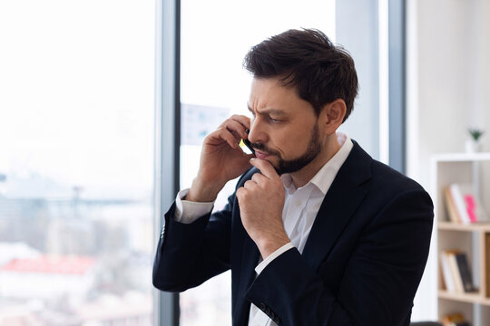 Young adult male in formal suit in office communicating on phone with contemplative expression.
