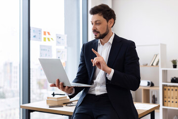 Naklejka premium Professional adult Caucasian man wearing suit seated at desk holding laptop explaining concept