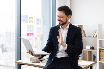 Businessman with beard in suit sitting in modern office having online conference waving, engaging in communication through laptop. Positive and professional workplace environment showcasing technology