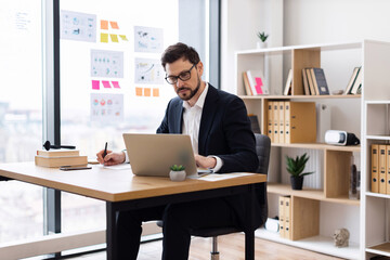 Naklejka premium Young adult male wearing formal attire working with laptop at office desk writing notes. Modern workspace includes documents, books and potted plants indicating productive and professional atmosphere
