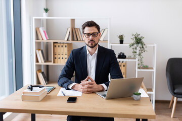 Confident adult male in suit working at desk with laptop and various supplies, professional office.