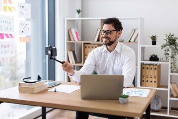 Young adult male recording video call, sitting by laptop in office environment, wearing glasses, business shirt, engaging with camera. Modern office setup with documents, tablet, and greenery