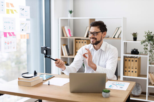 Young adult Caucasian male business professional creating video content in office setting. Posing professionally wearing glasses and formal white shirt. Modern workspace includes desk laptop notebooks
