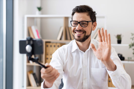 Middle-aged man recording video call with smartphone indoors expressing happiness and engagement. Dressed in white shirt, captured in bright modern office environment.