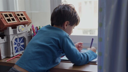 Young boy leaning on the windowsill, deeply focused on drawing, surrounded by colored pencils and toys. concentration and creativity of a child absorbed in an art activity-SD 480p