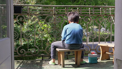 Young boy sitting on a small stool on a balcony, facing away, enjoying a quiet moment outdoors. the...