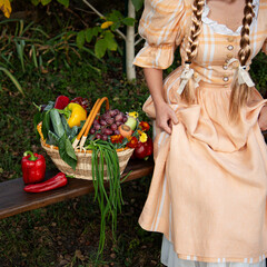 Harvesting lady in traditional outfit. Blondy with long braids, vegetable basket with grapes, green onion, red pepper pods, carrot. Cottage lifestyle, vintage Austrian dress in light peach color.