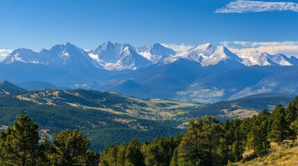 Obraz premium Wide mountain range view with snow-capped peaks, trees, and clear blue sky.
