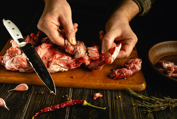A person is skillfully cutting and preparing fresh meat on a wooden board, surrounded by herbs, garlic, and spices. The kitchen has a warm, inviting atmosphere