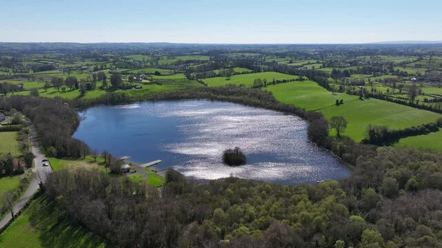 Hollywood Lake, County Monaghan, Ireland, April 2023. Drone panoramic orbit clockwise around Crannog in middle of shimmering rippled water reflecting sky and clouds above