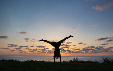 Silhouette slender girl standing on hands upside down at sunset on sea coast, concept of cheerfulness, activity, healthy lifestyle. young woman trains with pleasure. photo in motion. hello vacation