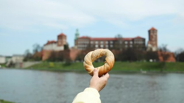 Hand holding a traditional Krakow bagel (obwarzanek) against the backdrop of the historic Wawel Castle and Vistula River. A perfect combination of local food, culture, and travel in Poland.