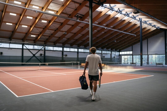 A man in sports clothes walks along the tennis court. He holds a bag of equipment and a tennis racket in his hands. Rear view, unrecognizable person