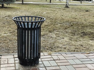 A black metal trash can in a spring park, next to a bench