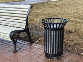 A black metal trash can in a spring park, next to a bench