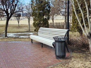 A black metal trash can in a spring park, next to a bench