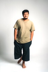 Fototapeta premium Portrait of an Asian Thai man with casual clothes standing barefoot on white background, showing relaxed attitude, natural confidence, cultural identity, and authentic lifestyle in studio shot