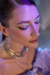 Naklejka premium In a creative indoor environment, a fashion model displays striking makeup featuring neon highlights on her eyelids. models elegant hairstyle and jewelry enhance the captivating look