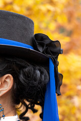 Black cylinder hat with blue ribbon and black bow (side view). Victorian fashion attributes, abstract, close-up. Retro lady, vintage walking outfit in blue color. Black-haired lady, hairstyle, curls. 