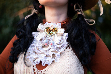 Handmade costume attribute, burlap and lacework jabot collar accessory. Black-hair, pigtails hairstyle. Retro fashion vibe, brown dress with white accents, straw hat fragment. Golden sequins heart bow