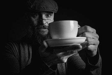 Black and white portrait, a mature bearded man in a stylish cap and glasses offers a cup of coffee with a mysterious smile. The spectacular lighting and dark background create a vintage mood.
