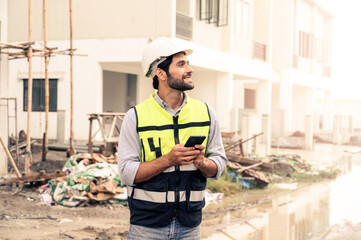 portrait of young engineer in vest with white helmet standing on construction site, smiling and holding smartphone for worker, internet, social media.