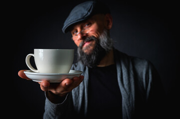 Mature bearded man with a stylish cap and glasses offers a cup of coffee with a mysterious smile....