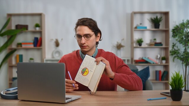 Young adult man spending time at home, man student sits at desk works on laptop, talking online by video call, writing in notebook, shows notes at screen.