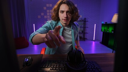 Young adult man spending time at home, man streamer gamer sits at desk looks at camera and pointing...
