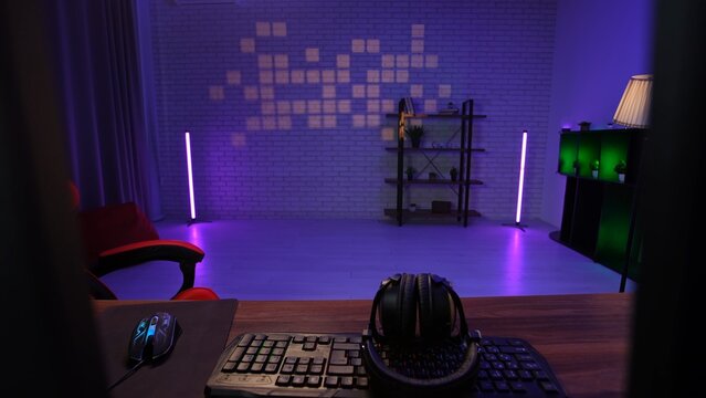 Young adult man home, shot of male living room with neon light, desk with computers, keyboard and gaming chair, modern streamer podcaster blogger room setup.
