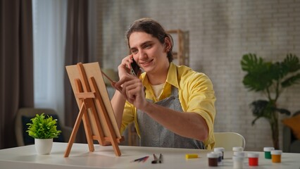 Young adult man spending time at home, man in apron sits at the table sketching drawing picture on portable easel and talking on smartphone.