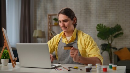 Young adult man spending time at home, man in apron sits at the table and buying art supplies...
