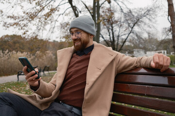Fototapeta premium A man enjoying the outdoors while using a smartphone, sitting on a park bench.