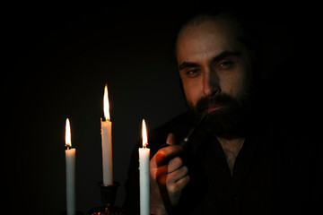 Man with contemplative expression holding a pipe, sitting in a dimly lit room with three burning...