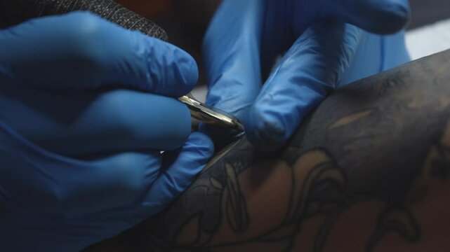 Close-up view of a tattoo artist in blue gloves creating a floral tattoo on a clients forearm. The tattoo machine is in motion as the artist outlines the design. Studio environment
