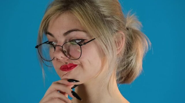 Disgruntled model with ponytail looks down rubbing chin. Serious young woman purses lips expressing dissatisfaction on blue background slow motion