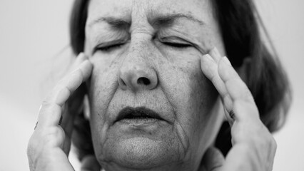 Elderly woman pressing fingers to her temples, eyes squeezed shut, showing visible signs of intense...