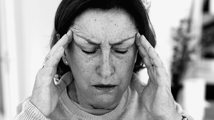 Elderly woman struggling with severe headache, massaging temples with visible concentration. Facial...