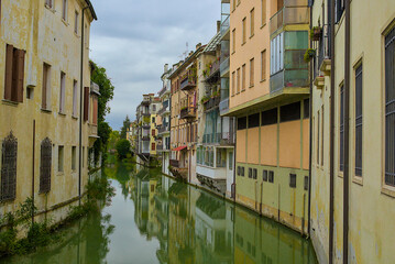 Narrow waterways glide between ancient buildings adorned with plants in Padua, Italy. Every window and balcony tells a timeless story of the city, muted reflections shimmering under overcast skies.
