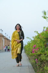 Fototapeta premium A graceful young woman wearing a peach and orange floral salwar kameez, adorned with traditional jewelry, poses elegantly on a sunlit pathway surrounded by lush greenery