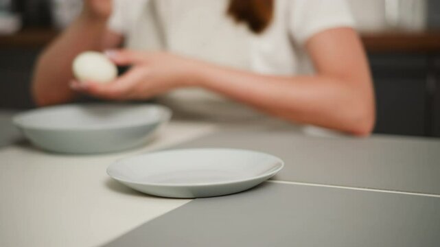 Slightly blurred view of woman collecting peeled egg from dish and returning it while using bowl to clean hand, with two plates on kitchen table and part of apron visible
