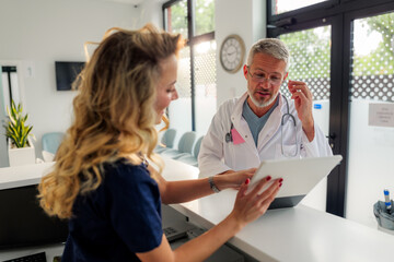 Senior doctor presenting medical information on a tablet while collaborating with a young nurse at the reception desk in a bright and clean hospital waiting room